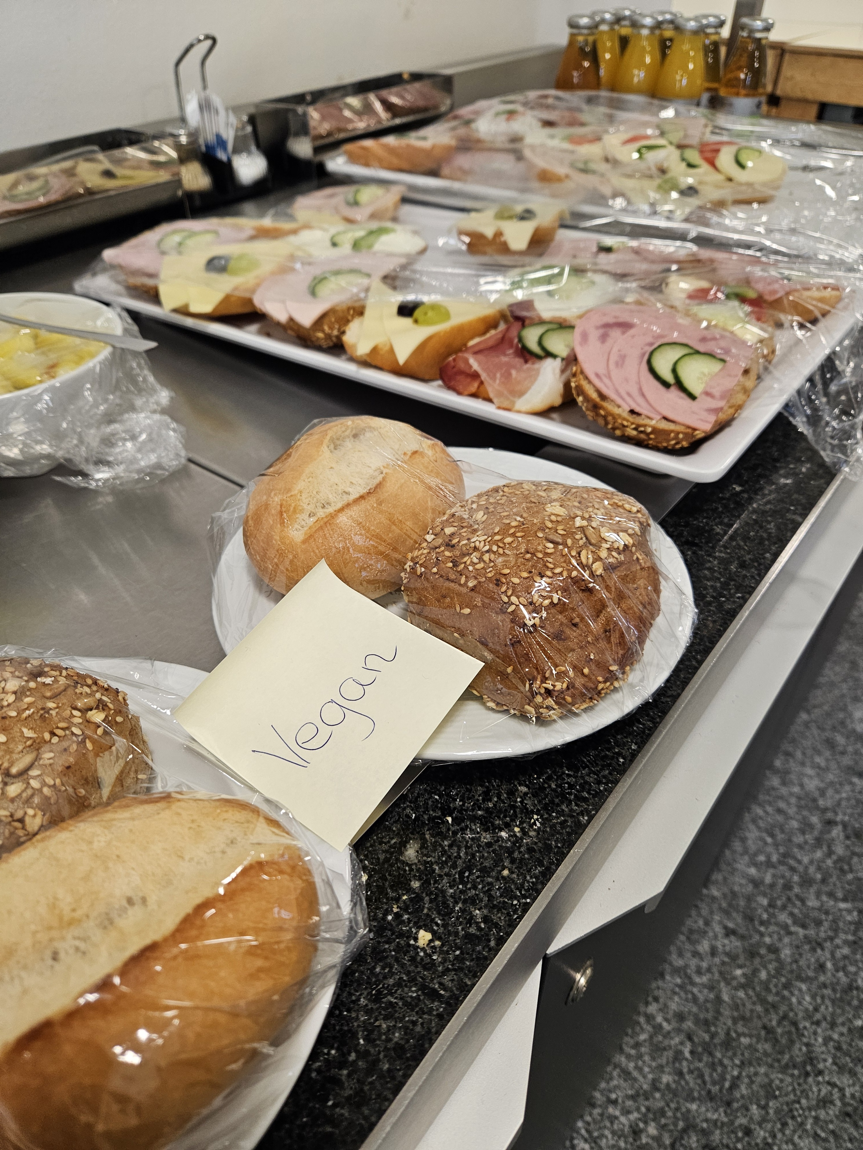 Buffet mit belegten Brötchen (Tierwurst und Kuhkäse). daneben 2 Teller mit je 2 unaufgeschnittenen Brötchen und einem Aufkleber mit der Aufschrift "vegan"