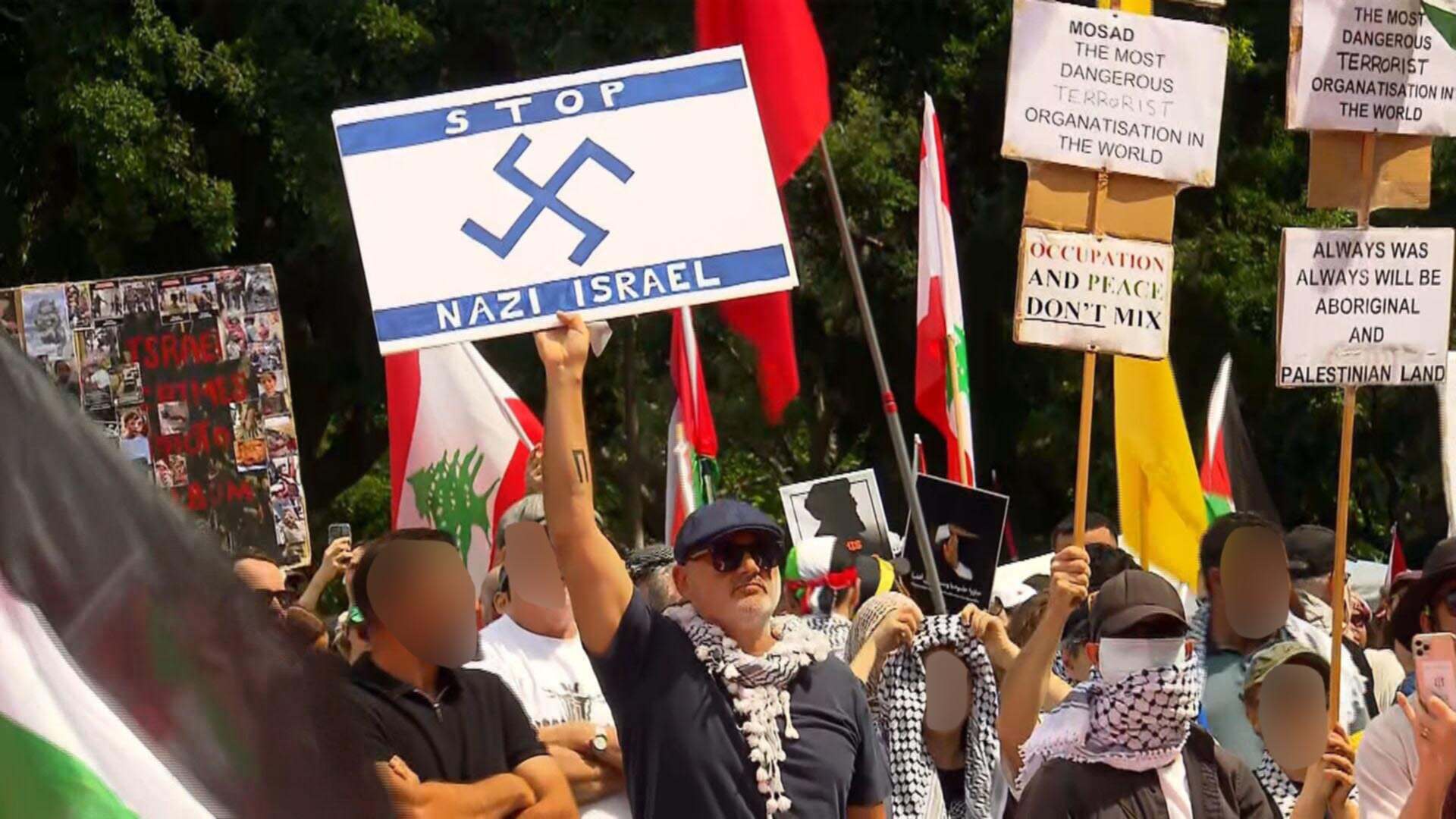 A man in a protest crowd holding up a hand-painted placard in the style of an Israeli flag with a swastika replacing the star of david, and the words "STOP NAZI ISRAEL"