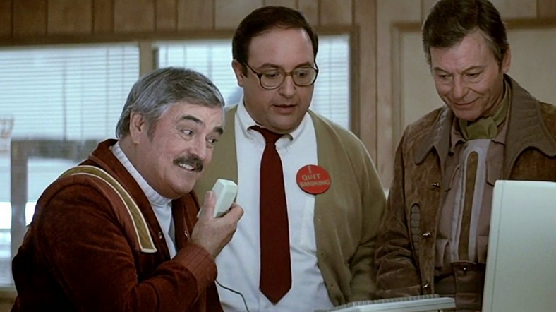 screencap of the "Star Trek IV: The Voyage Home" scene at Plexicorp where Montgomery Scott (James Doohan) is sitting in front of an Apple Macintosh and holding its mouse in his hand as if it where a microphone, while Dr. Nichols (Alex Henteloff) and Dr. Leonard "Bones" McCoy (DeForest Kelley) stand next to him.