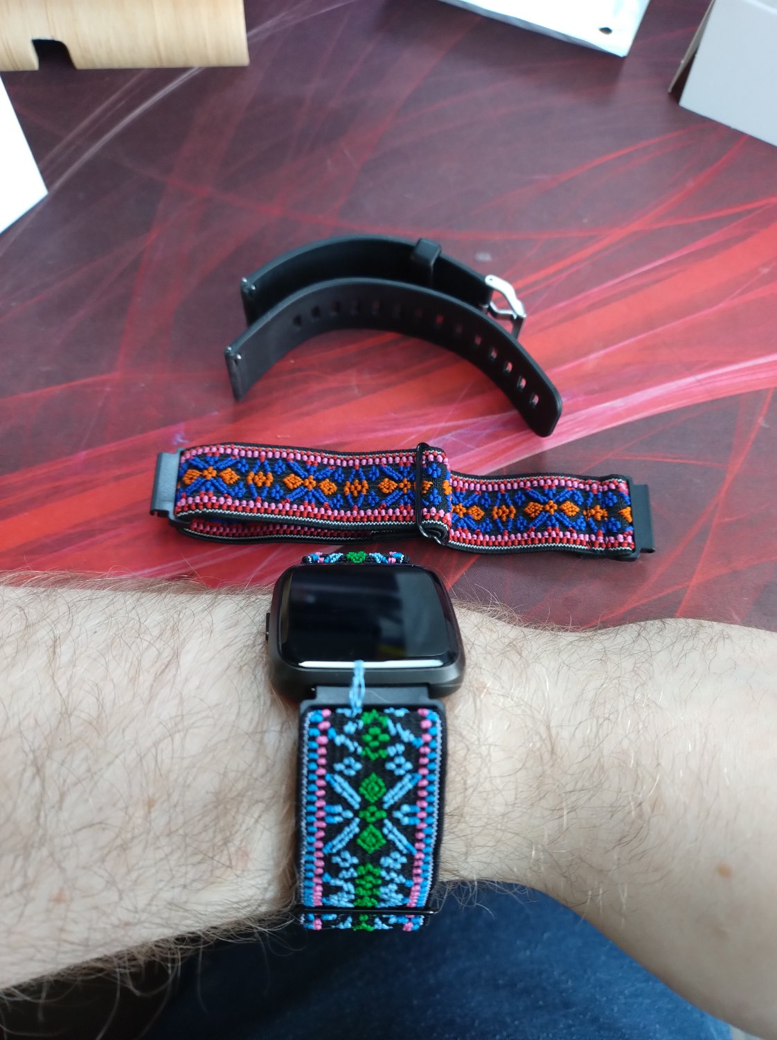 A sleeping PineTime smart watch with a green, light blue, blue, and pink watch band holding it onto his pale arm. Above is a secondary band in the colours, blue, orange, red, and pink, above them is the black rubber bands that came with the pinetime.