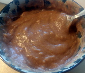 Bowl of cooked sorghum porridge. Reddish-brown colour, a bit lumpy but otherwise smooth. Glistening in the light. Eating spoon sitting in it.