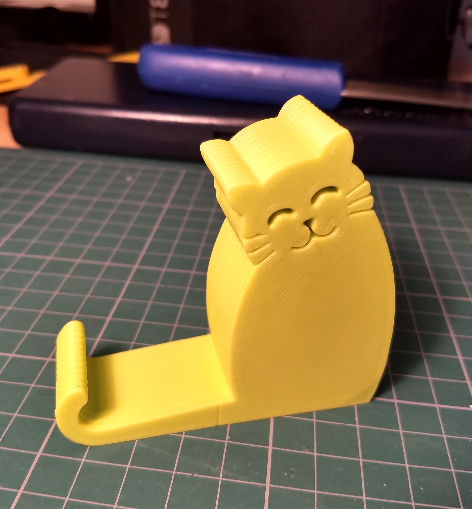 A stand for a smartphone in the stylized shape of a cat, with the cat tail curling up to retain the bottom of the phone. The part is 3D printed in light green colour.