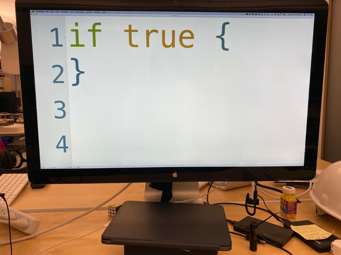 photograph of a Macintosh computer. a programming text editor with line numbers enabled fills the screen. its font size is so huge that there is only room for four lines of code. the first two lines are "if true {" and "}". lines 3 and 4 are blank. a 3x3 rubik's cube and various other objects are on the desk.