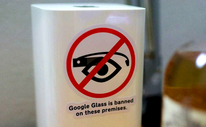 a sign with a red circle and line through a drawing of an eyeball with a google glass in front of it, above the text "Google Glass is banned on these premises"