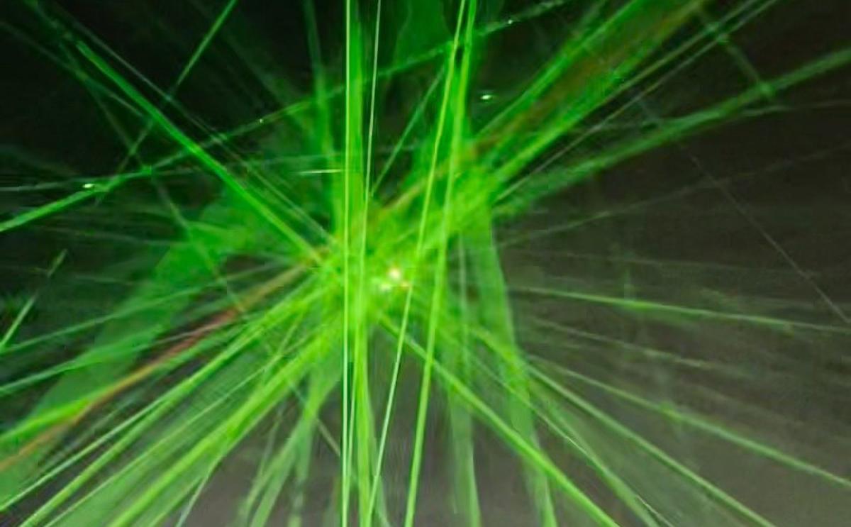 Hundreds of green lasers shine up at an airplane against the night sky